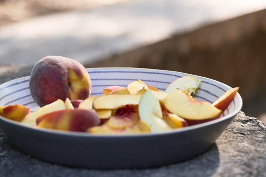 Close-up of a striped ceramic serving dish filled with sliced peaches and apples, with one whole peach and a softly blurred outdoor background.