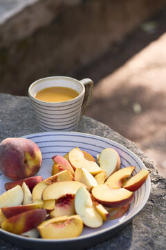 Close vertical view of sliced peaches and apples served in a striped ceramic dish with a cup of orange juice on a rustic outdoor stone table.