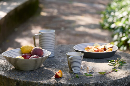Coordinated striped ceramic bowl, pitcher and cup displayed with fresh peaches, apples and lemon on a rustic outdoor stone table in soft dappled sunlight.