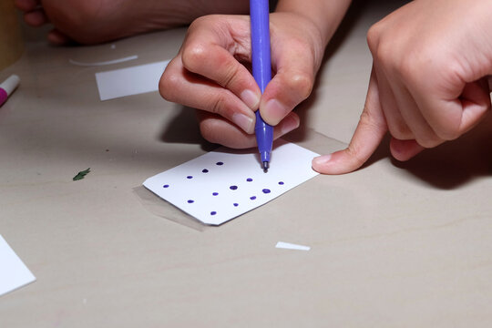 person's hands carefully drawing purple dots on a small white rectangular piece of paper using a marker pen.