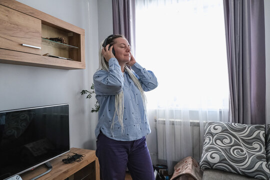 A young Caucasian woman with long blonde hair enjoys music while wearing headphones in a cozy living room. A television and a sofa are visible in the background.