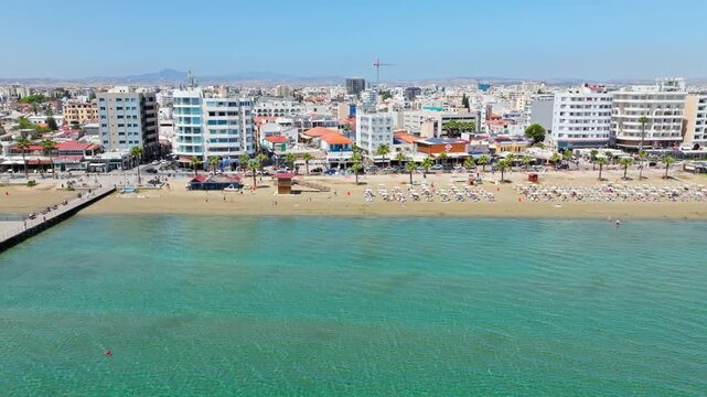 Drone view of Larnaca seafront in Cyprus. Calm shallow water, sandy beach, promenade and waterfront buildings create a peaceful coastal city view.