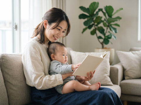 ソファに腰掛け絵本を眺める我が子を見つめる母親の女性