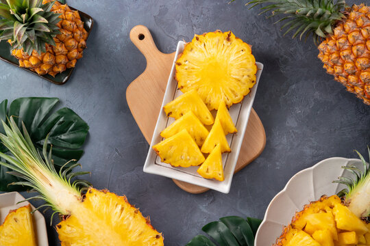 Fresh sliced pineapple fruits with green leaves on wooden board over dark gray textured background.