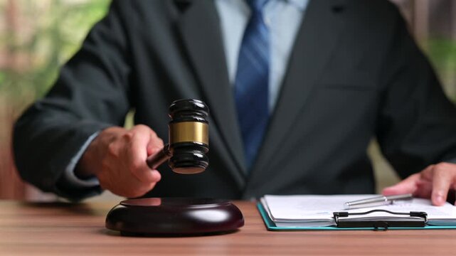 Male judge wearing dark suit holds wooden gavel over sound block near clipboard containing paper documents, representing justice, legal trial, court decision, authority, auction bidding process.