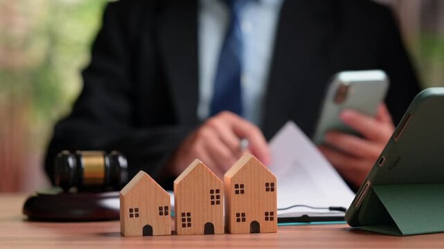 Lawyer wearing suit holding mobile phone sits behind wooden house models beside legal gavel, tablet device representing real estate auction, property dispute, mortgage foreclosure proceedings.