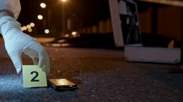 Forensic investigator placing evidence marker at crime scene, detective collecting smartphone evidence at night, police investigation with yellow number tag