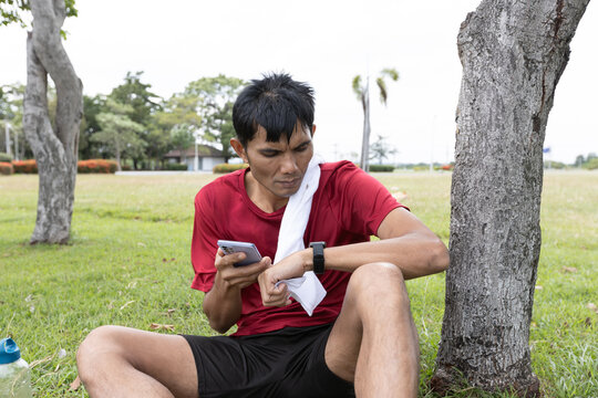 Asian male athlete reviewing fitness performance data on smartwatch and smartphone after workout in an outdoor park, representing fitness tracking, recovery, and digital fitness lifestyle.