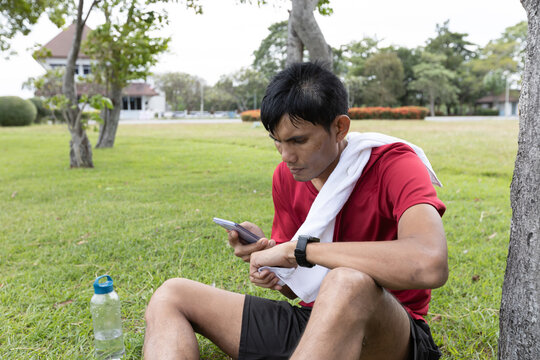 Asian male athlete reviewing fitness performance data on smartphone and smartwatch after workout in an outdoor park, representing fitness tracking, recovery, and digital fitness lifestyle.