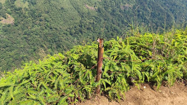 Rusty barbed wire fence post along a mountain cliff edge overlooking a green forest valley.