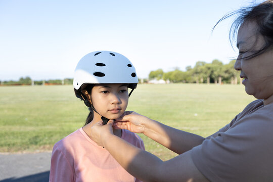 Close up of an Asian girl wearing a safety helmet while a parent adjusts the strap outdoors, showing child safety, parental care, and protection.