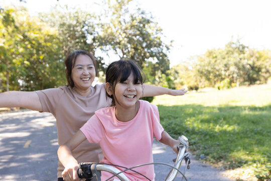 Asian mother and daughter cycling outdoors with arms outstretched and laughing together, showing playful family interaction and joyful lifestyle moment.