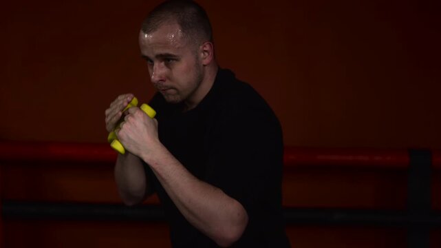 Male boxer in black shirt holds bright yellow hand weights in guarded stance inside boxing ring, with red ropes, faint ghosting, dark warm backdrop, and focused training energy