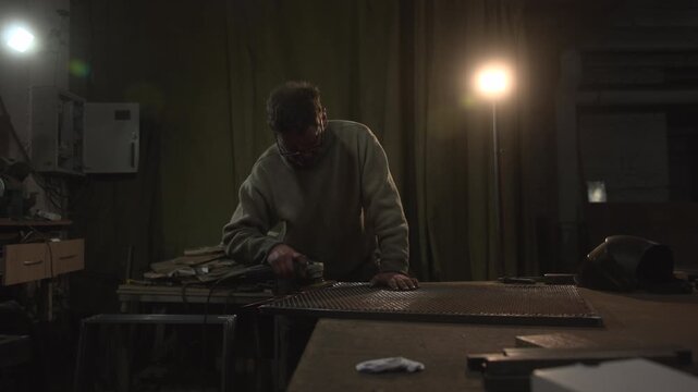 Man using handheld grinder on metal sheet inside dim workshop with bright sparks, warm backlight, rough industrial textures, tools, cabinets, curtains, helmet, and scattered materials around workbench