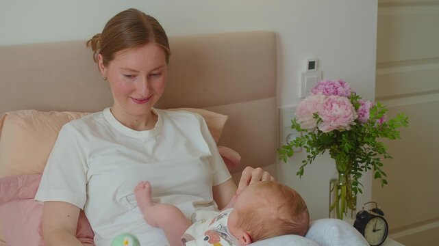 Mother places rattle into infant hand during quiet play on bed. Guided grasping introduces object handling, tactile awareness, hand control, trust, and patient maternal assistance.