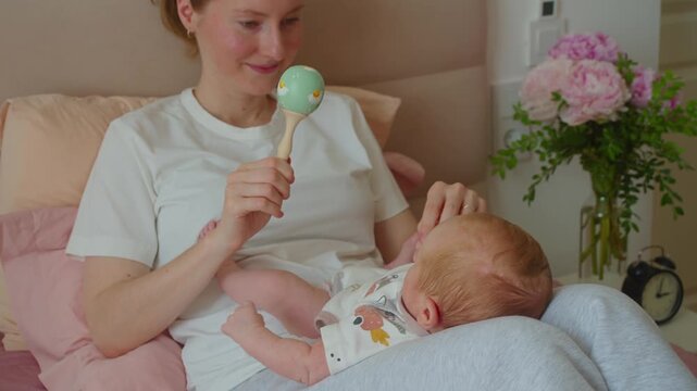 Cinematic shot follows mother picking up rattle before play with newborn on bed. Visual transition highlights preparation, maternal attention, anticipation, and warm family interaction.