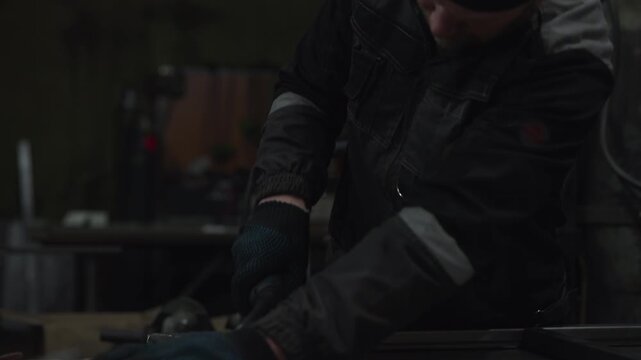 Focused worker in dark protective jacket and blue gloves leaning over metal equipment inside low light industrial workshop with bright sparks and blurred machinery