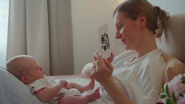 Mother shows black-and-white card while infant follows moving image on bed. Visual tracking supports focus, early perception, sensory learning, curiosity, and attentive parent guided play.