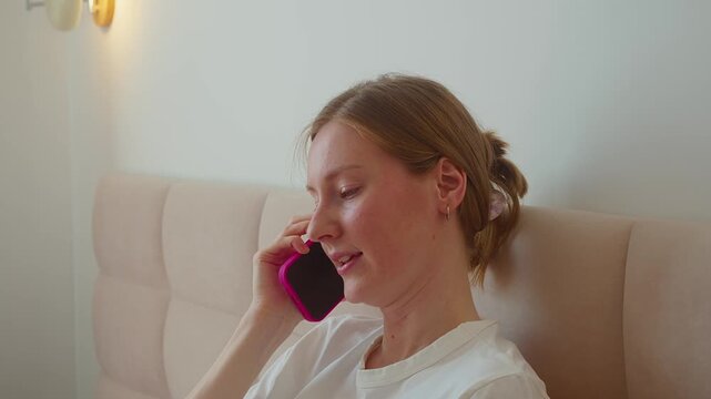 Smiling mother talks on smartphone while baby rests nearby on bed. Positive expression adds warmth, relaxed communication, maternal presence, emotional comfort, closeness, and everyday family care.