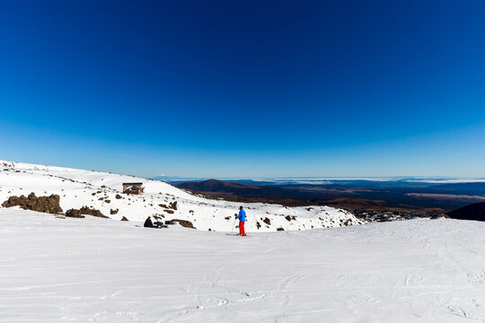 A skier stands on the snowy Whakapapa slopes under a clear blue sky, with distant mountain ranges and rocky terrain stretching toward the horizon