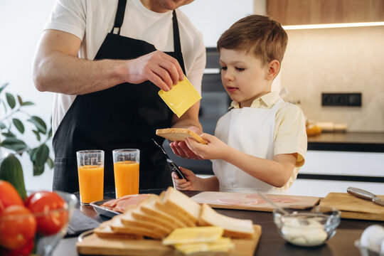 Adult male and young boy preparing sandwiches together in a modern kitchen, with fresh ingredients and drinks on the counter, showcasing a family cooking moment