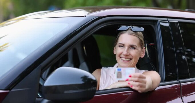 Smiling woman sitting in the car and showing her new driving license out of window to the camera