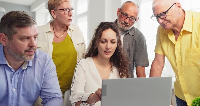 Instructor teaches seniors students using laptop technology. Group stands around computer in classroom as woman guides them and answers questions. Technology training for senior students.