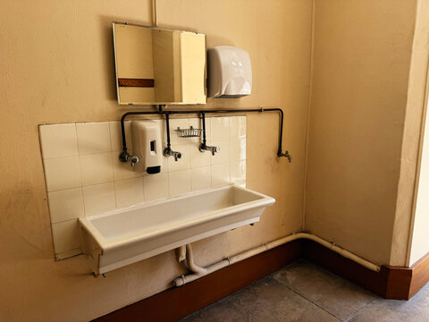 Vintage communal sink and soap dispensers in a tiled restroom corner in Paris, France with mirror, exposed pipes and warm plaster walls