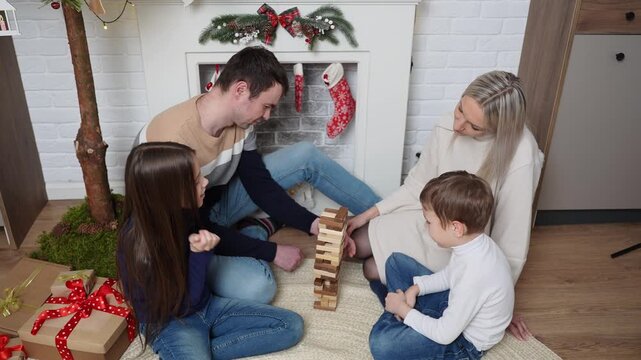 Happy family playing board game at Christmas, parents with kids stacking wooden blocks tower on floor near decorated fireplace, cozy winter holiday home activity, table game concept