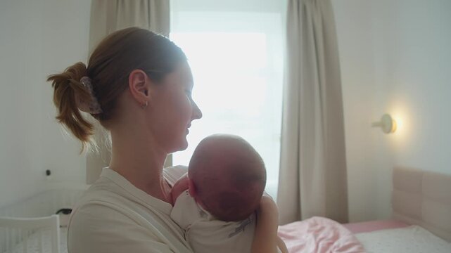 Mother carries crying baby across bedroom, keeping child close against her body. Movement becomes part of soothing effort, combining safe support, quick response, and steady maternal presence.