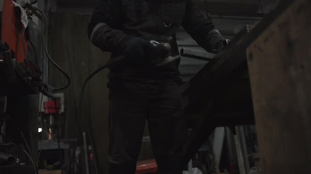 Worker in dark protective workwear uses handheld grinder on metal inside dim industrial workshop, sending bright orange sparks across rough surfaces with strong diagonal motion and focused technical