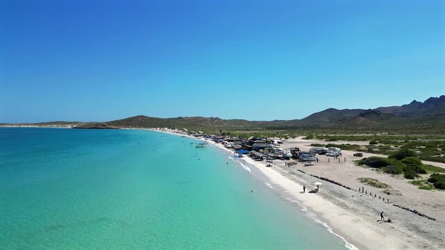 Static drone shot looking down the long stretch of El Tecolote Beach near La Paz, with a large beachfront restaurant and gathering place visible where crowds have gathered along the shore.