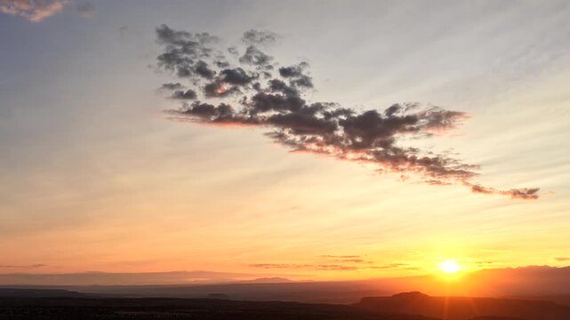 4K drone timelapse of a glowing golden sunrise over a dark mountain and canyon landscape. A single sparse, textured cloud formation shifts smoothly through a soft pastel sky at dawn.