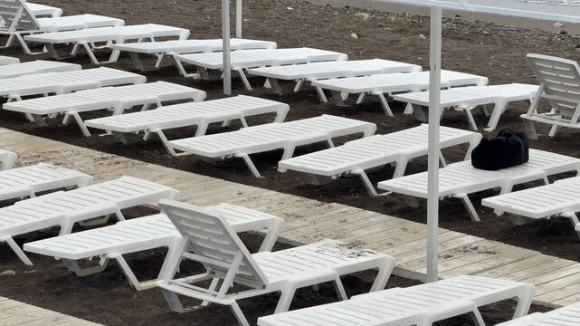 Empty white beach sun loungers arranged in rows. Horizontal panning shot of a serene coastline scene.