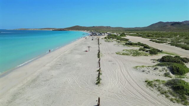 Drone flies high above a wooden fence line at El Tecolote Beach, looking toward the larger eastern stretch of the beach with faint hints of people scattered beneath umbrellas, near cars, and around a