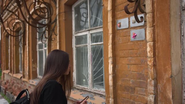 Young woman lowers smartphone beside weathered orange brick facade while inspecting memorial plaques, old window frames, wrought iron scrollwork, and peeling paint in daylight