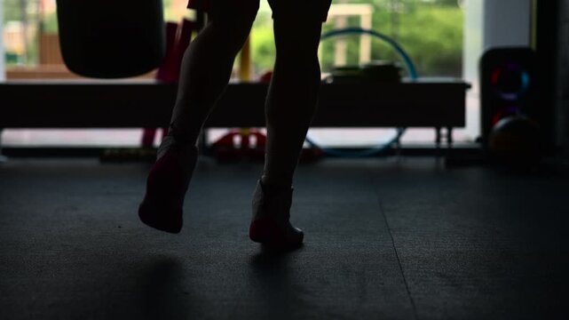 Silhouetted lower legs and feet rising onto toes above dark gym floor inside moody fitness space, backlit by bright window with blurred equipment and calm focused training mood