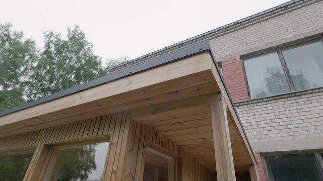 Low-angle exterior view of a small wooden modular cabin with a deep roof overhang, timber posts, dark metal trim, and trees beside an industrial building