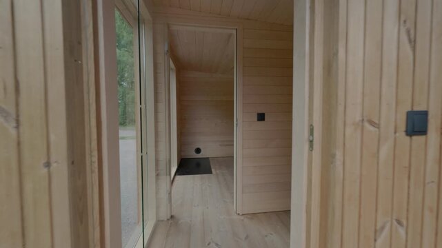 Front entrance of a compact modular timber cabin with a covered porch, glass side panel, open doorway, and natural wood cladding beside a rural road