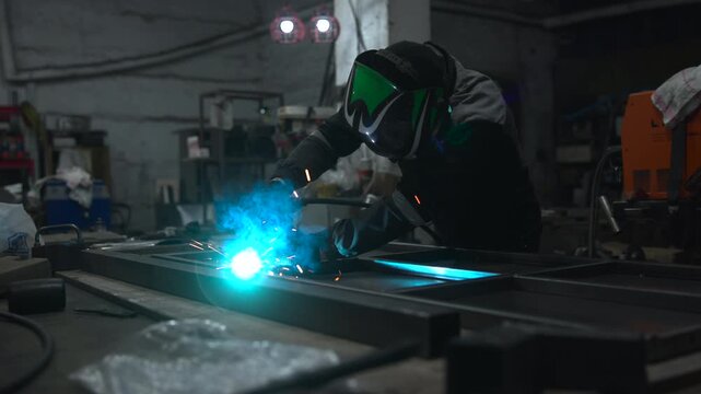 Skilled worker bends over steel panel during bright welding action inside dim industrial workshop with heavy machinery, glowing sparks, dark protective clothing, gritty metal surfaces, focused work