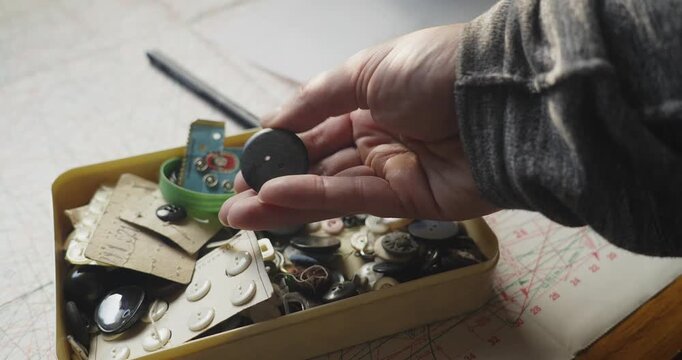 Close-up of a dressmaker wearing a wrist pincushion while selecting buttons from a vintage button box, highlighting tailoring, garment finishing and craftsmanship.