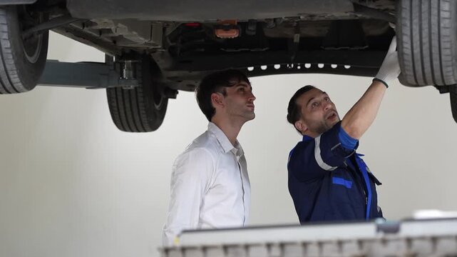 Car mechanic customer garage repair lift vehicle inspection undercarriage tire service worker pointing auto part clean workshop