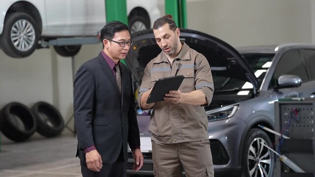 Mechanic customer reviewing car inspection on tablet in garage, auto service consultation and repair beside vehicle open hood