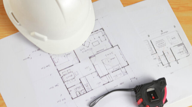 White hard hat and laser distance meter placed on architectural blueprints, representing construction planning, civil engineering, workplace safety, building design, and site inspection.