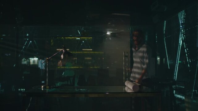 Focused man in white shirt bends over tabletop with hands on desk, studying bright sheet beside rolled papers inside moody industrial workspace with dark surfaces, reflective glass, neon green