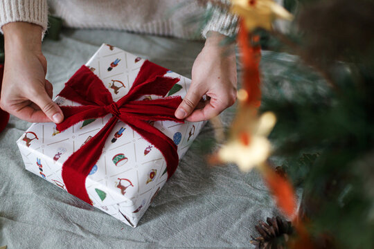 Wrapping stylish xmas presents. Woman in cozy sweater wrapping Christmas gift box with red ribbon at home. Festive DIY holiday preparation, top view. Merry Christmas