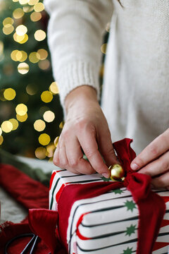 Wrapping stylish xmas presents. Woman in cozy sweater wrapping Christmas gift box with red ribbon at home. Festive DIY holiday preparation near decorated xmas tree with golden bokeh lights