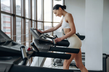 Fototapeta premium Female fitness enthusiast in white sportswear adjusts treadmill settings while exercising in modern gym with large windows and fitness equipment in the background