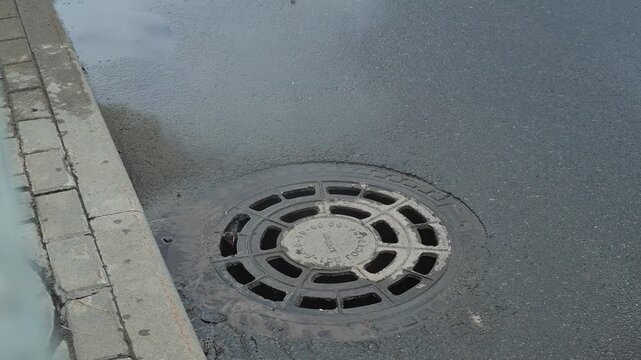 A metal drain cover is placed on a wet street, surrounded by a concrete edge.