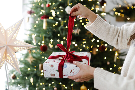Merry Christmas. Hands in cozy sweater opening a red bow on Christmas gift box. Festive holiday celebration near decorated xmas tree with warm golden bokeh garland lights at home